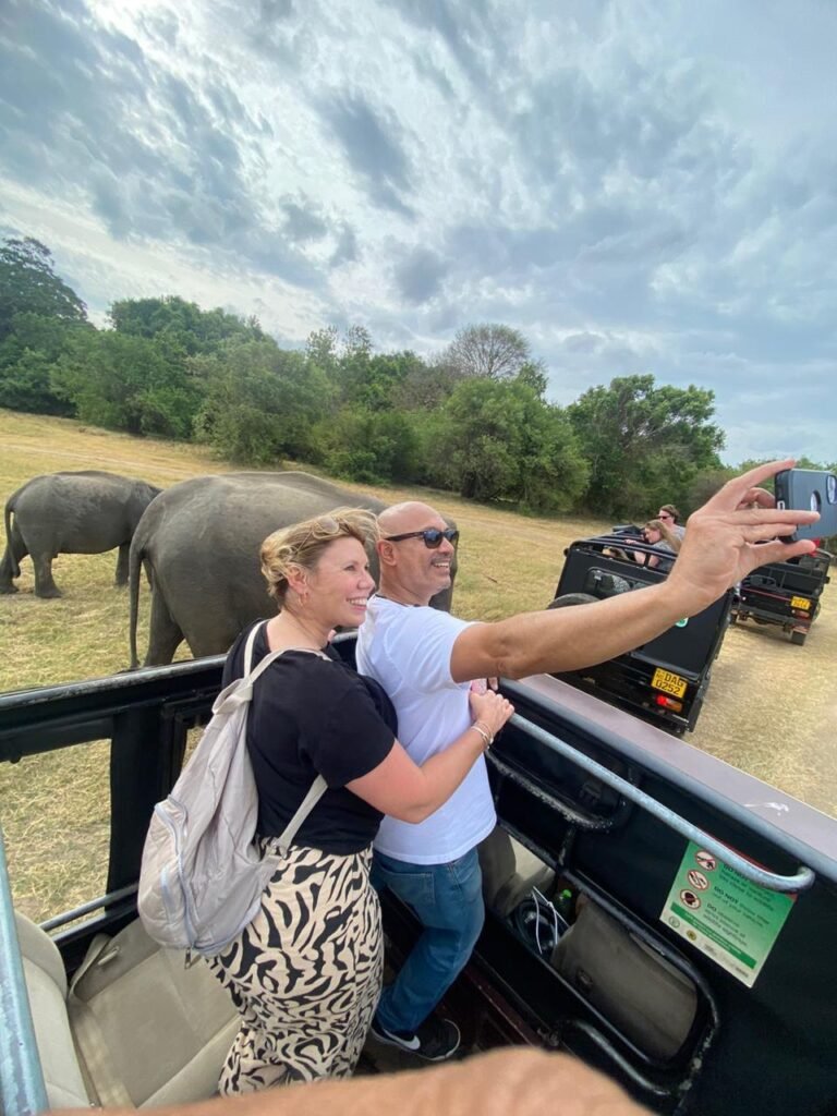 In this unforgettable shot from Udawalawe National Park, the joy of safari exploration is vividly captured. The image shows a group of safari-goers, their faces alight with excitement as they observe a family of majestic elephants in their natural habitat. Seated in an open-top jeep, the visitors are treated to a breathtaking view of several elephants gracefully roaming the golden savannah. The expansive landscape and distant hills provide a stunning backdrop, enhancing the sense of adventure and connection with nature. This moment perfectly embodies the thrill of wildlife encounters and the shared wonder of experiencing the untamed beauty of Sri Lanka.